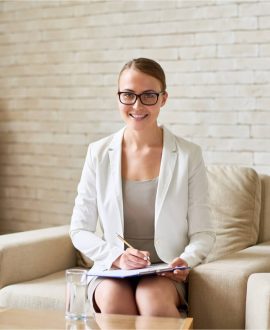A woman in glasses and a white blazer sits on a beige armchair, holding a clipboard and pen, smiling at the camera. A glass of water is on the table in front of her.