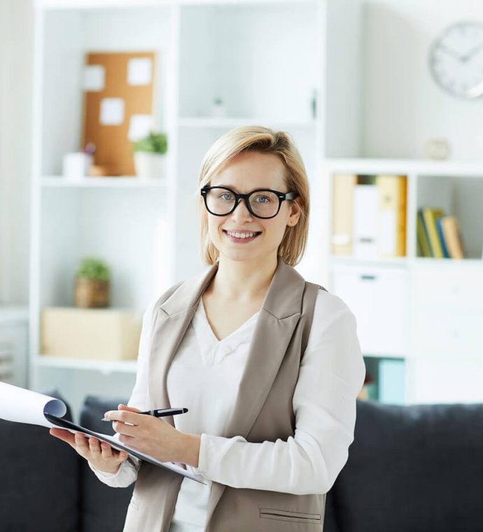 Person with glasses holding a clipboard stands in an office with shelves and a couch in the background.