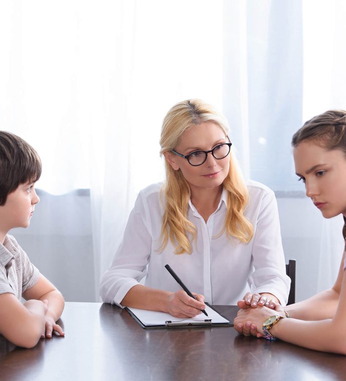 A woman with glasses sits at a table with a young boy and a teenage girl, taking notes on a clipboard.
