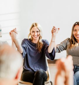 Three women seated in a circle hold hands and smile during a group meeting.