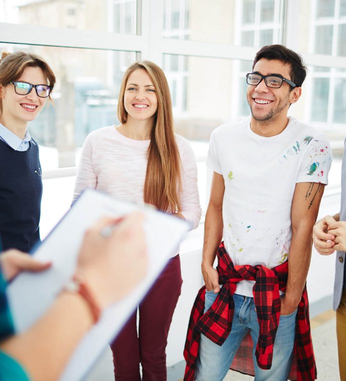A group of four people standing and smiling while one person in the foreground writes on a clipboard. They are indoors with large windows in the background.