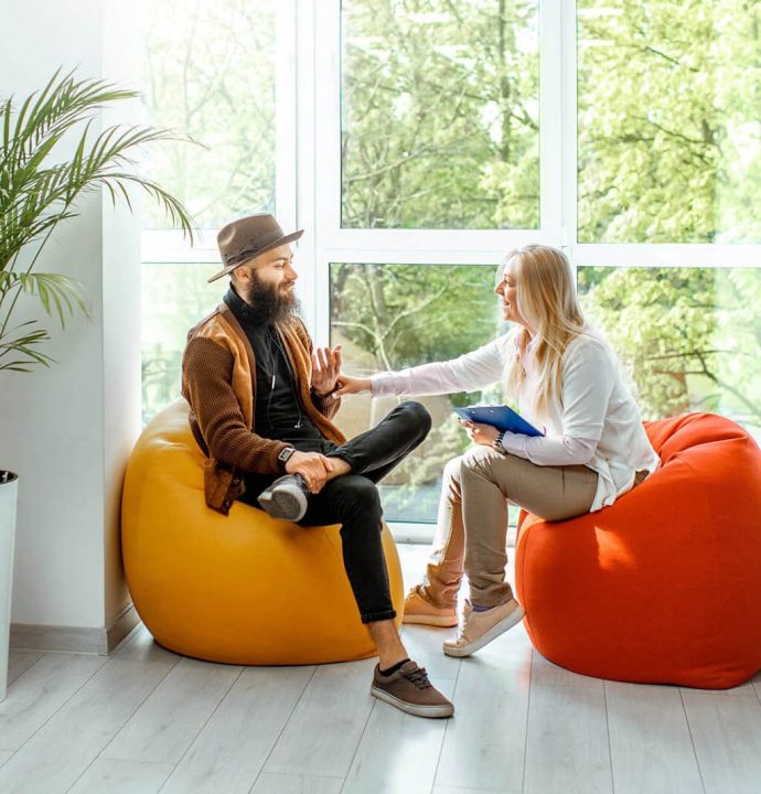 Two people sit on colorful bean bags near a large window with greenery outside. One holds a clipboard, and they appear to be engaged in conversation. A potted plant is on the left. Two people sit on colorful bean bags near a large window with greenery outside. One holds a clipboard, and they appear to be engaged in conversation. A potted plant is on the left.