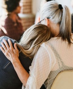 Two individuals sitting, with one person resting their head on the other's shoulder. The person on the right has gray hair and wears a white blouse.