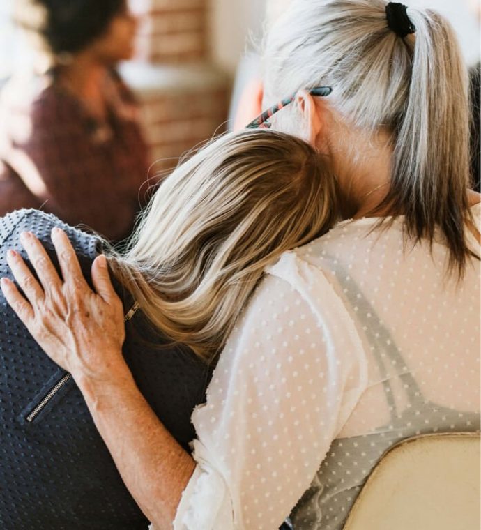 Two individuals sitting, with one person resting their head on the other's shoulder. The person on the right has gray hair and wears a white blouse.