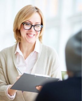 A woman with glasses smiles while holding a clipboard, engaging with another person. A woman with glasses smiles while holding a clipboard, engaging with another person.