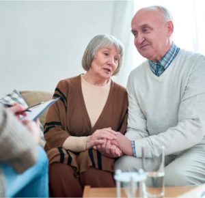An elderly couple sits on a couch holding hands, talking with a person taking notes in the foreground. Glasses of water are on the table.