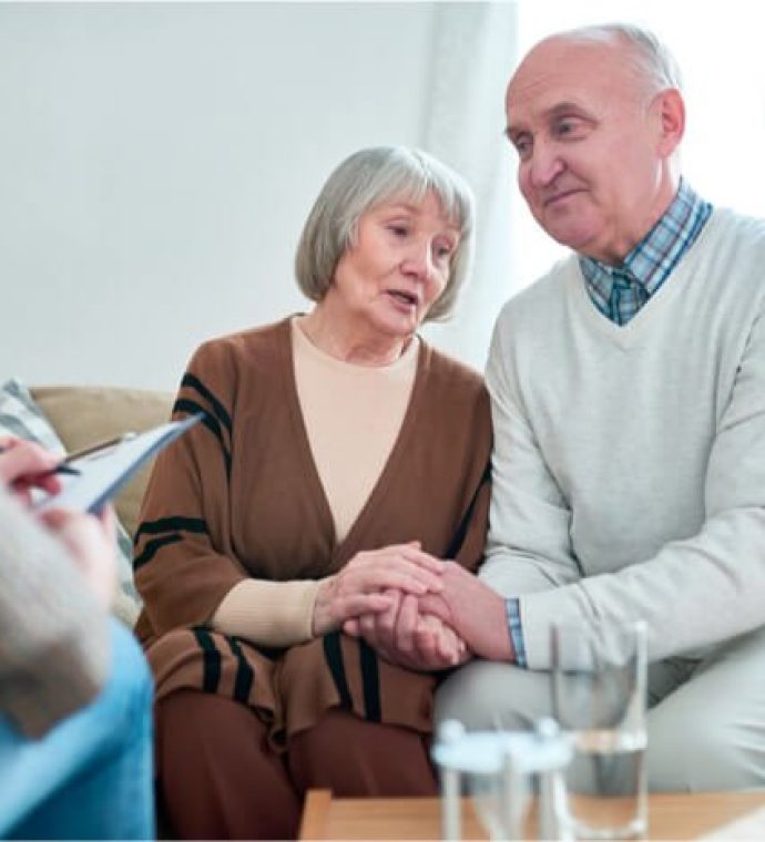 An elderly couple sits on a couch holding hands, talking with a person taking notes in the foreground. Glasses of water are on the table.