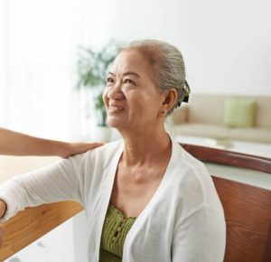 An elderly woman with gray hair smiles while sitting at a table. A person's hand rests on her shoulder. The background includes a plant and a couch.